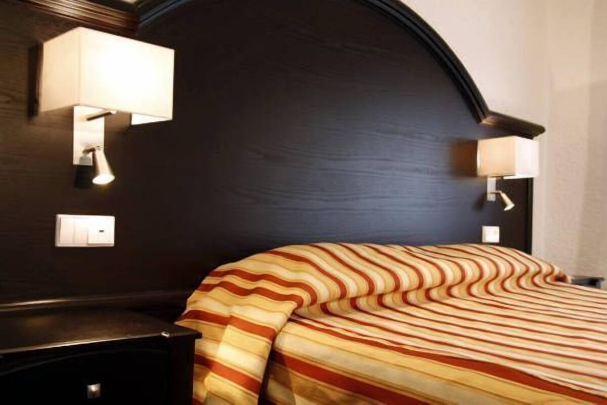 A hotel room with a dark wooden headboard, two wall-mounted lamps, a bed with a striped blanket, and a nightstand with a lamp.