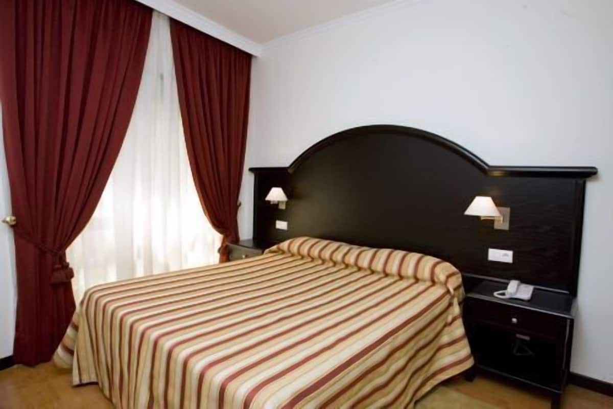 A hotel room with a bed, a nightstand, a lamp, and a window with curtains.