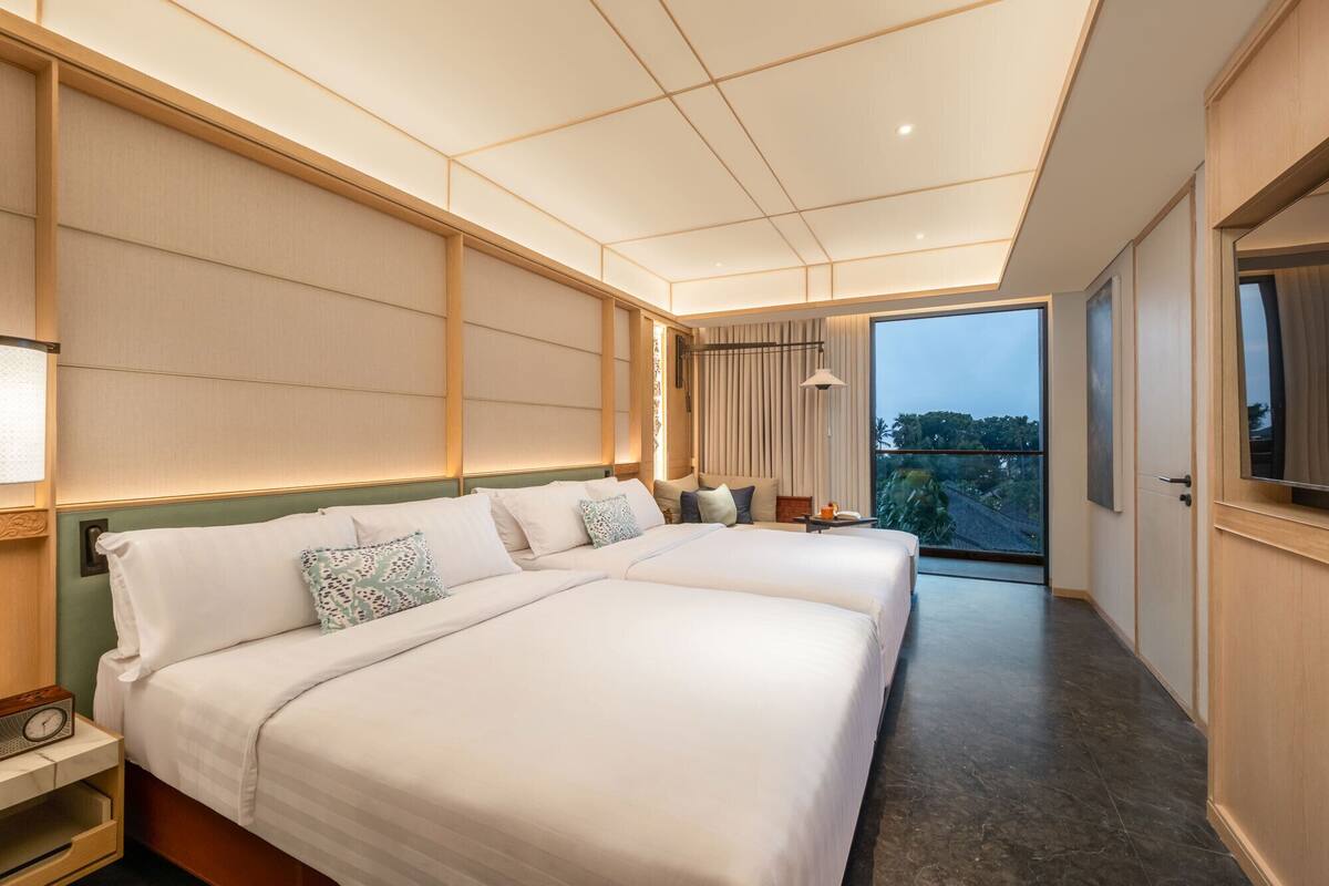 A modern hotel room with a large bed, bedside tables, a view through a glass door, and a ceiling with a geometric pattern.