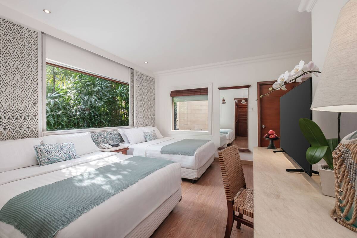 A modern hotel room with two beds, a large window, a desk, and a view of greenery.