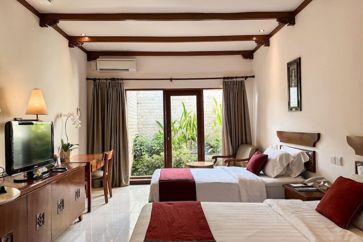 A hotel room with two beds, a TV, a desk, and a view of greenery through the window.