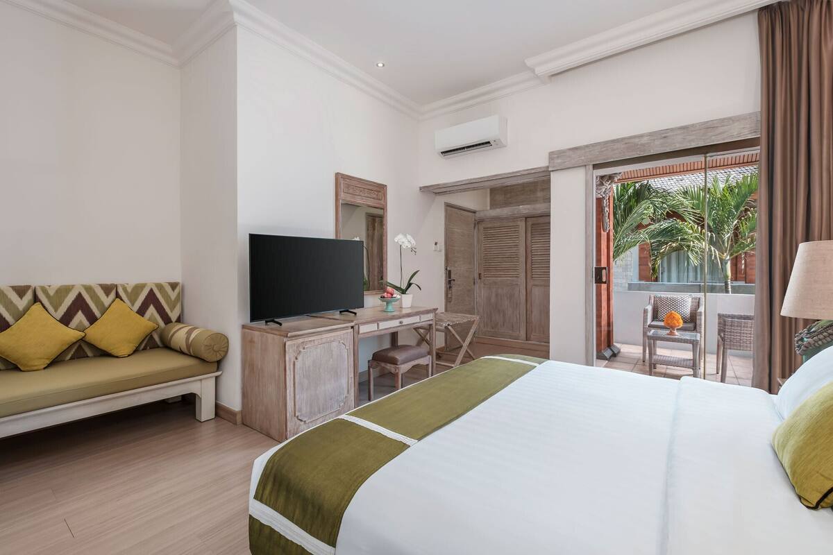 A hotel room with a bed, a TV, a desk, a sofa, and a balcony with plants.
