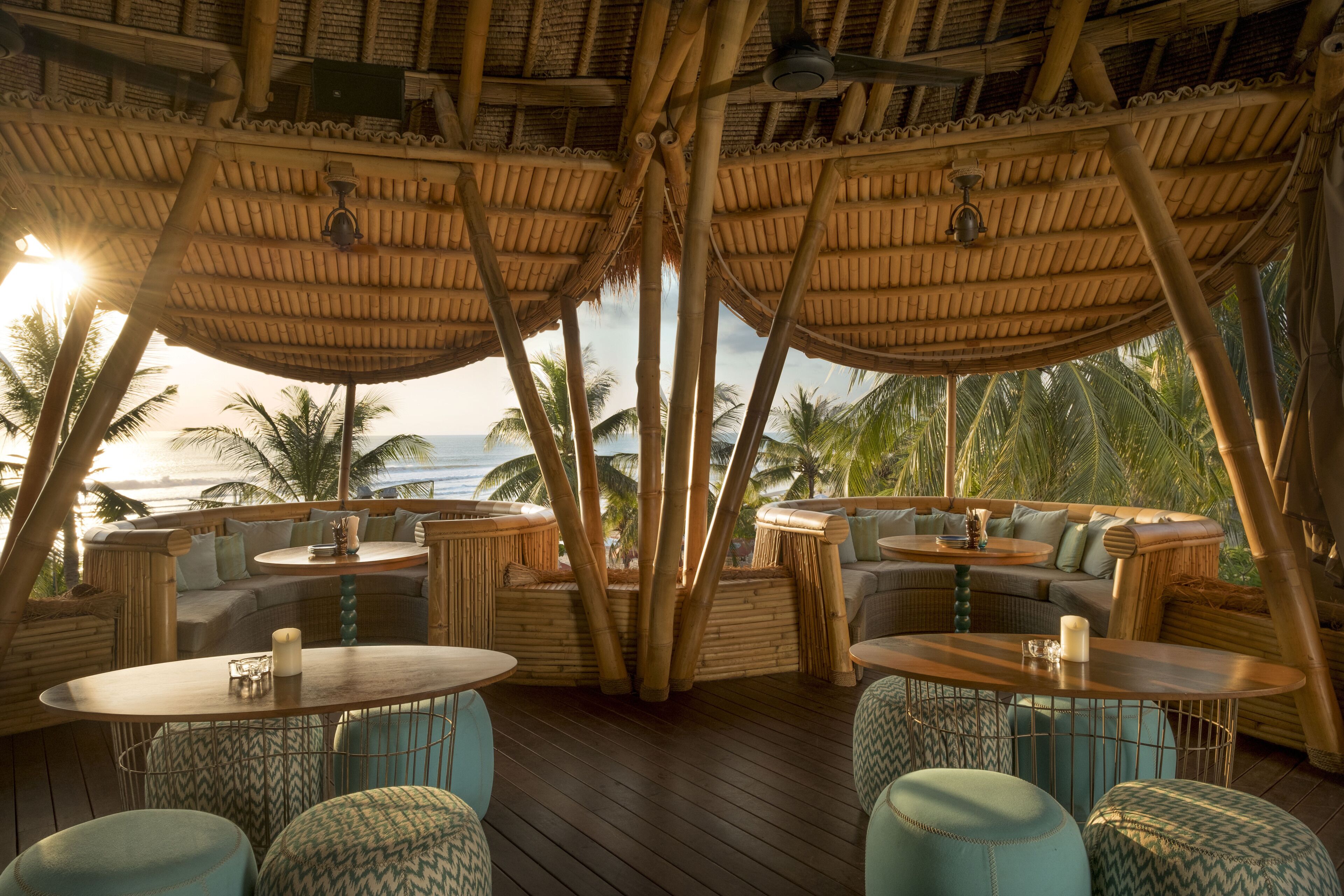 An outdoor lounge area with bamboo furniture, round tables, and a view of the ocean.