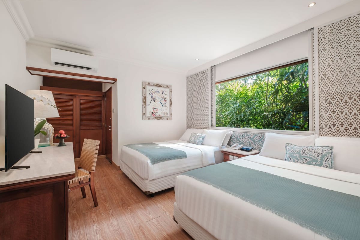 A bedroom with two beds, a desk, a chair, and a window with a view of greenery.