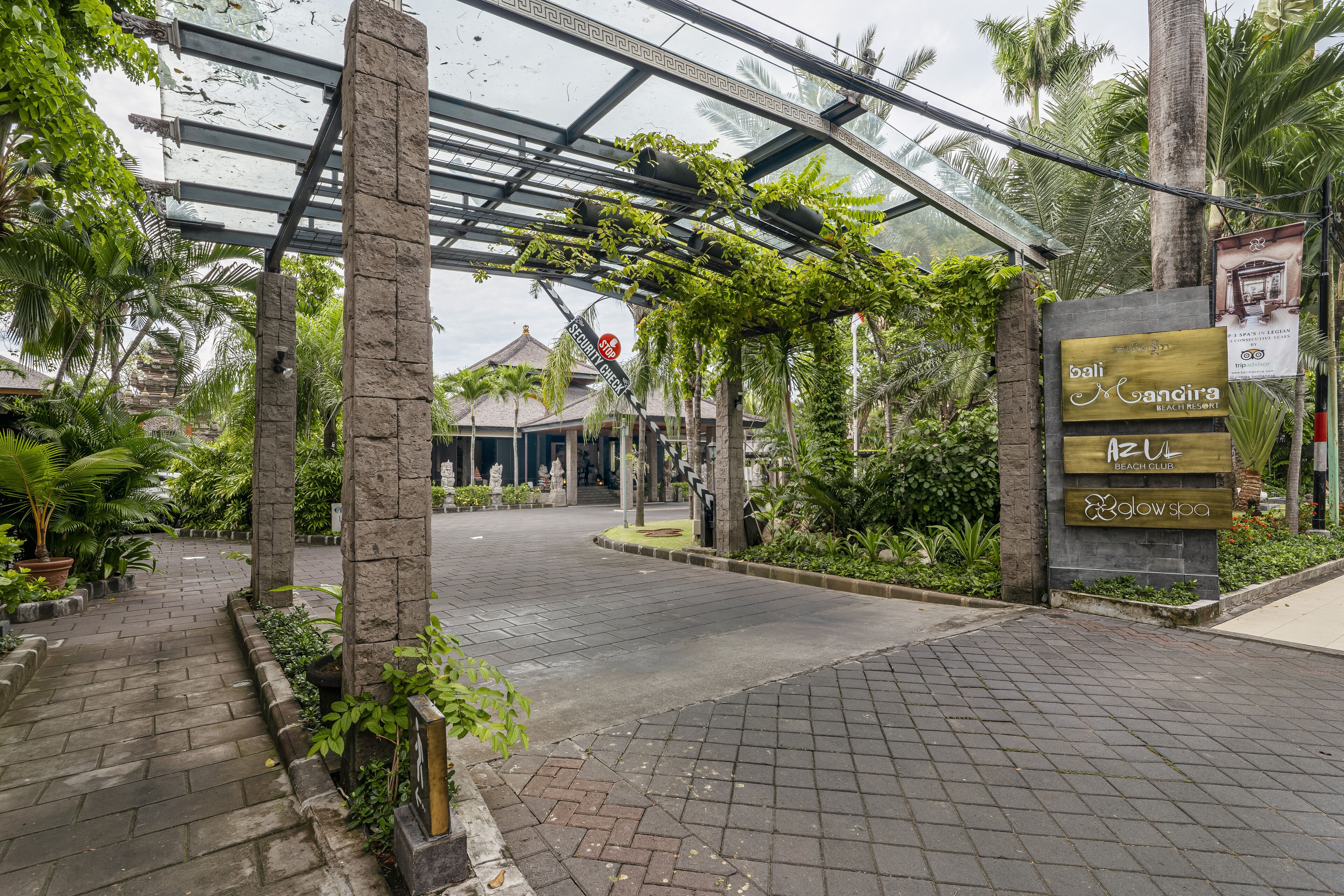 L'entrée d'un complexe hôtelier tropical, avec une allée couverte, des palmiers et un panneau indiquant « Bali Mandira Beach Club ».