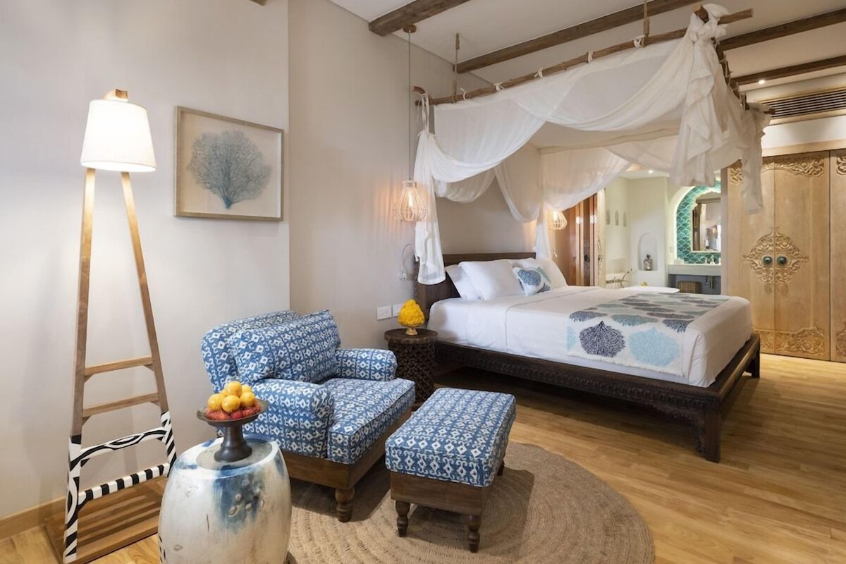 A bedroom with a canopy bed, a side table with a lamp, a chair, and a patterned ottoman.