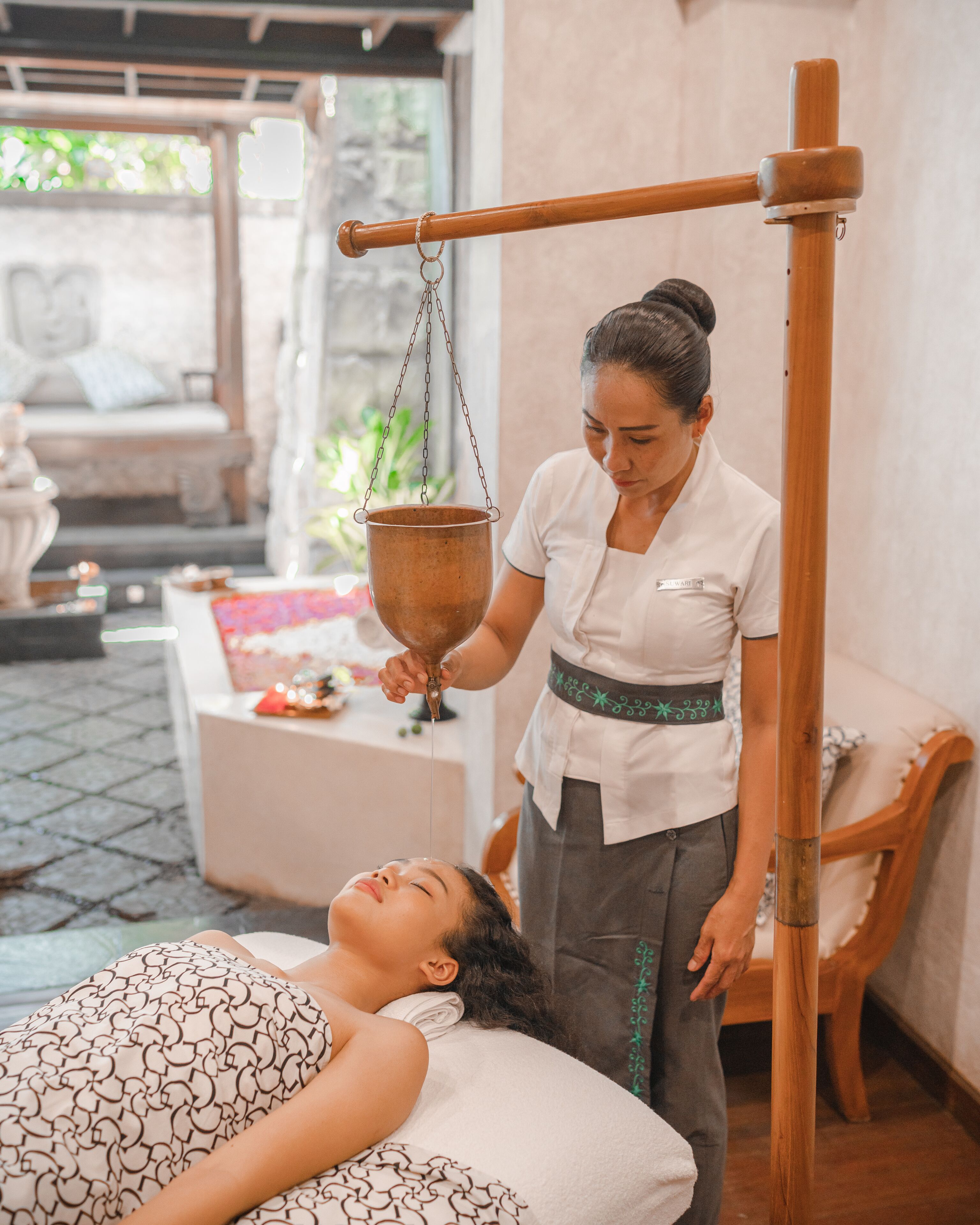 Eine Person erhält eine traditionelle Massage in einem Raum mit einem hölzernen Massagetisch, einem hängenden Kupfergefäß und einem Steinfußboden.
