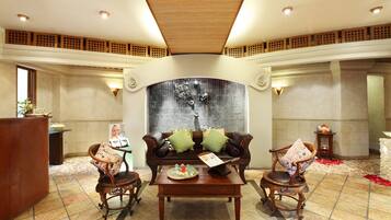 Un lobby amplio con techo alto, un área de estar central con sofá y sillones, un mostrador de recepción y una pared decorativa.