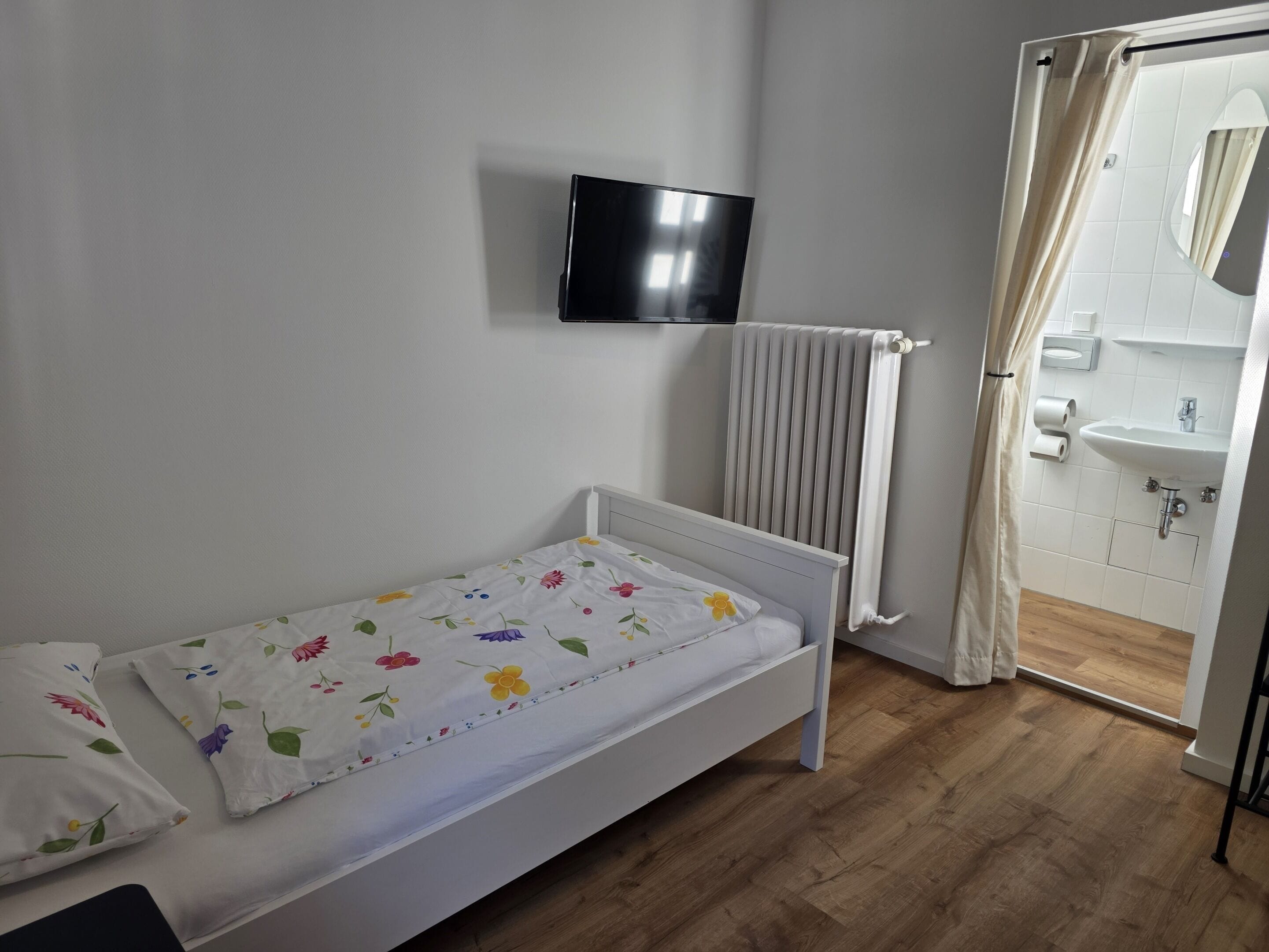 A small, single-bed room with a white bedspread, a mounted TV, a radiator, and a bathroom visible through an open door.