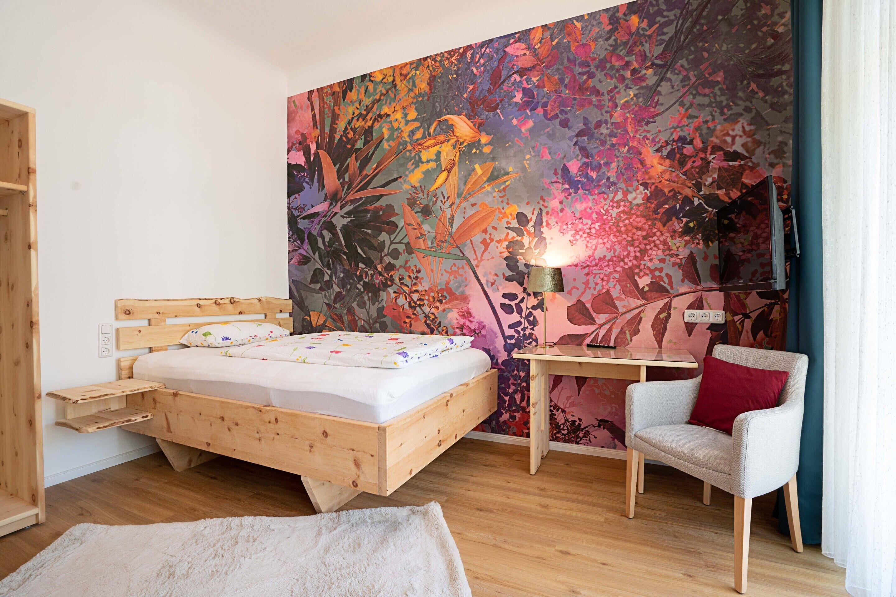 Une chambre avec un lit en bois, un bureau, une chaise et un mur décoré d’une fresque florale aux couleurs vives.