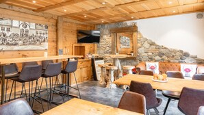 A wooden interior with a long dining table, bar stools, and a stone fireplace. - Hotel Alpina (Leukerbad)