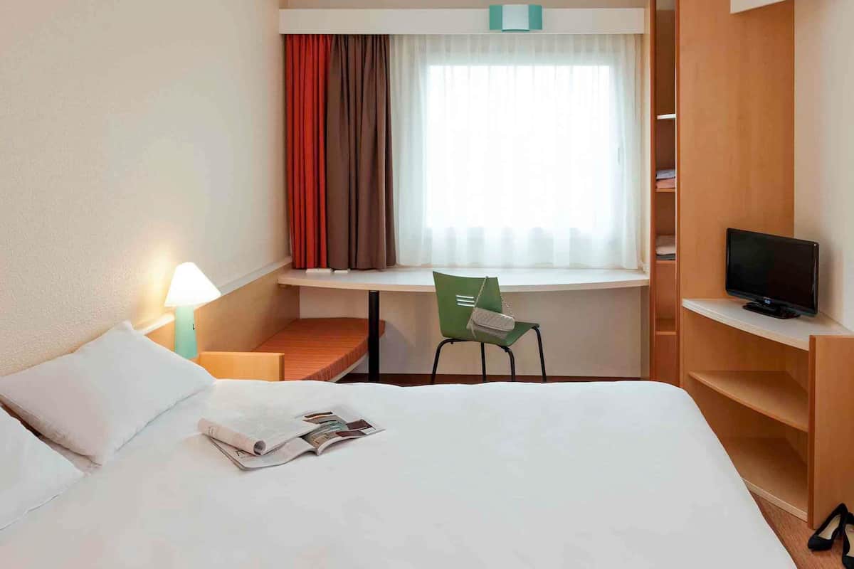 Une chambre d’hôtel comprenant un lit, un bureau avec une chaise, une télévision et une fenêtre avec des rideaux.