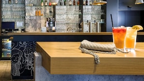 A bar area with a wooden counter, two drinks, a handbag, and a wall with shelves of bottles and glasses. - ibis Berlin City Potsdamer Platz (Berlin)