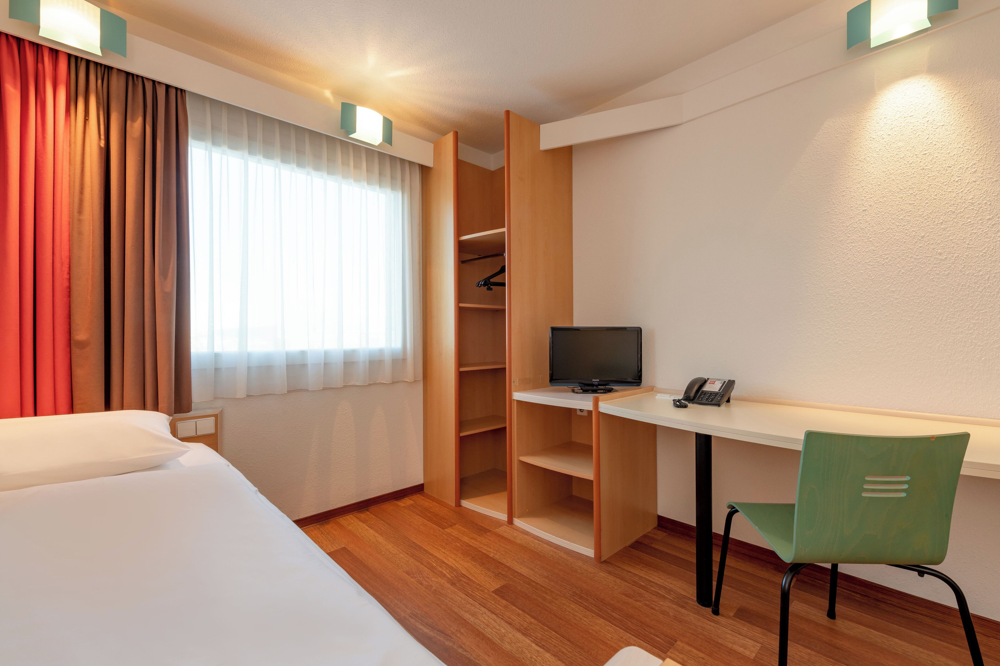 A hotel room with a bed, a desk with a computer, a chair, a wardrobe, and a telephone.