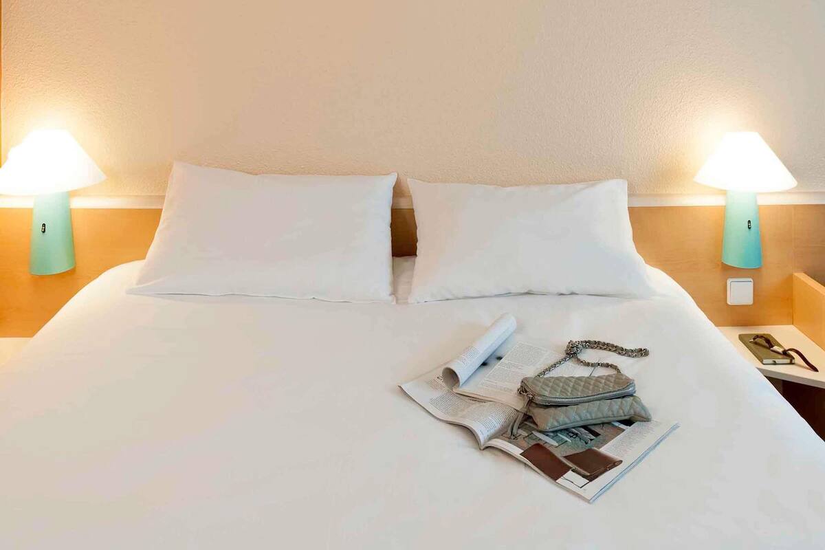 Un lit bien fait, avec du linge de lit blanc, deux lampes de chevet et une petite table sur laquelle se trouvent un livre et un sac à main.
