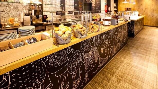 A breakfast bar with baskets of pastries, a coffee station, and a wall with artistic designs. - ibis Berlin City Potsdamer Platz (Berlin)
