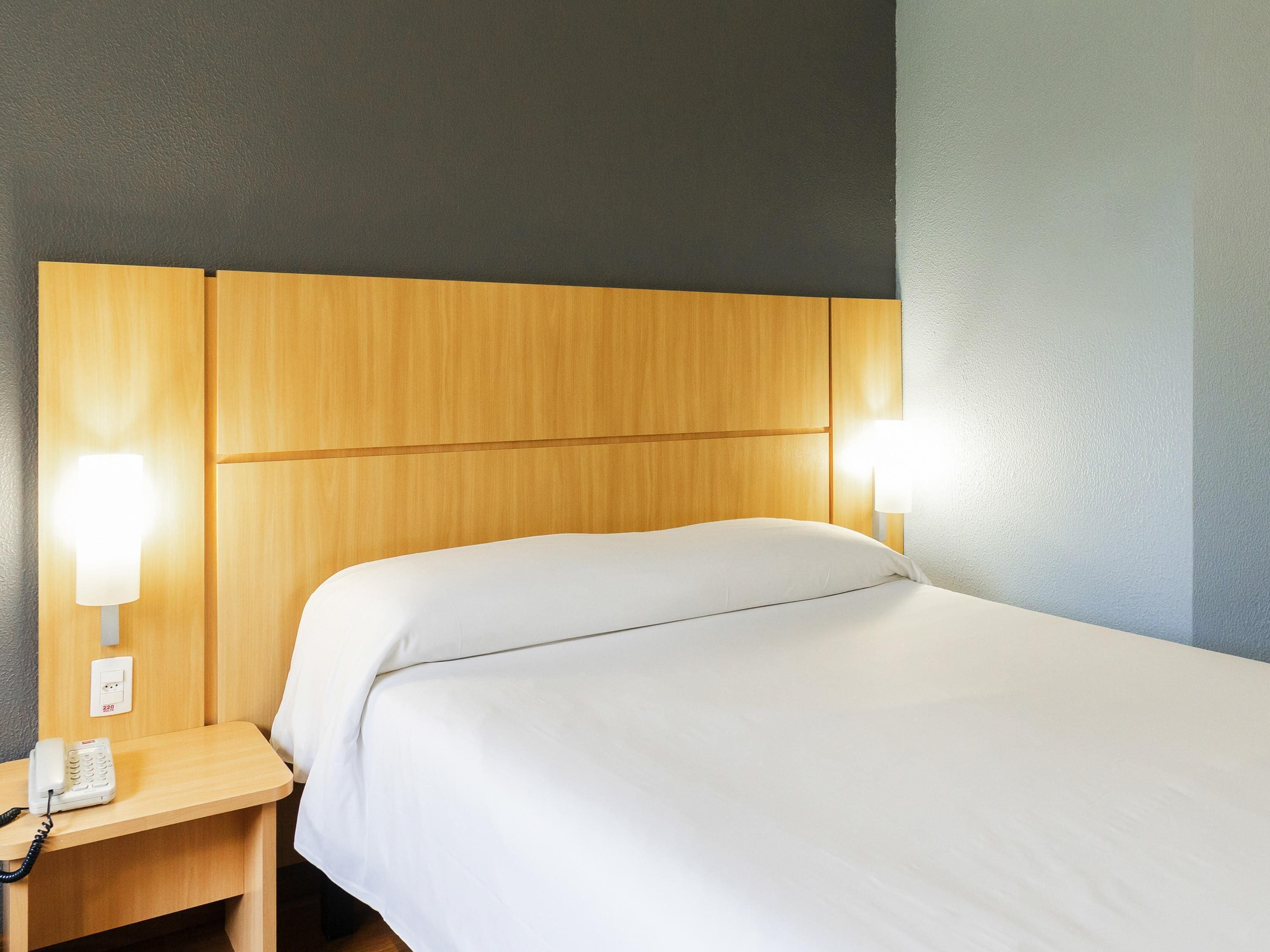 Quarto de hotel com cabeceira de madeira, mesa de cabeceira com telefone e duas luminárias fixadas na parede.