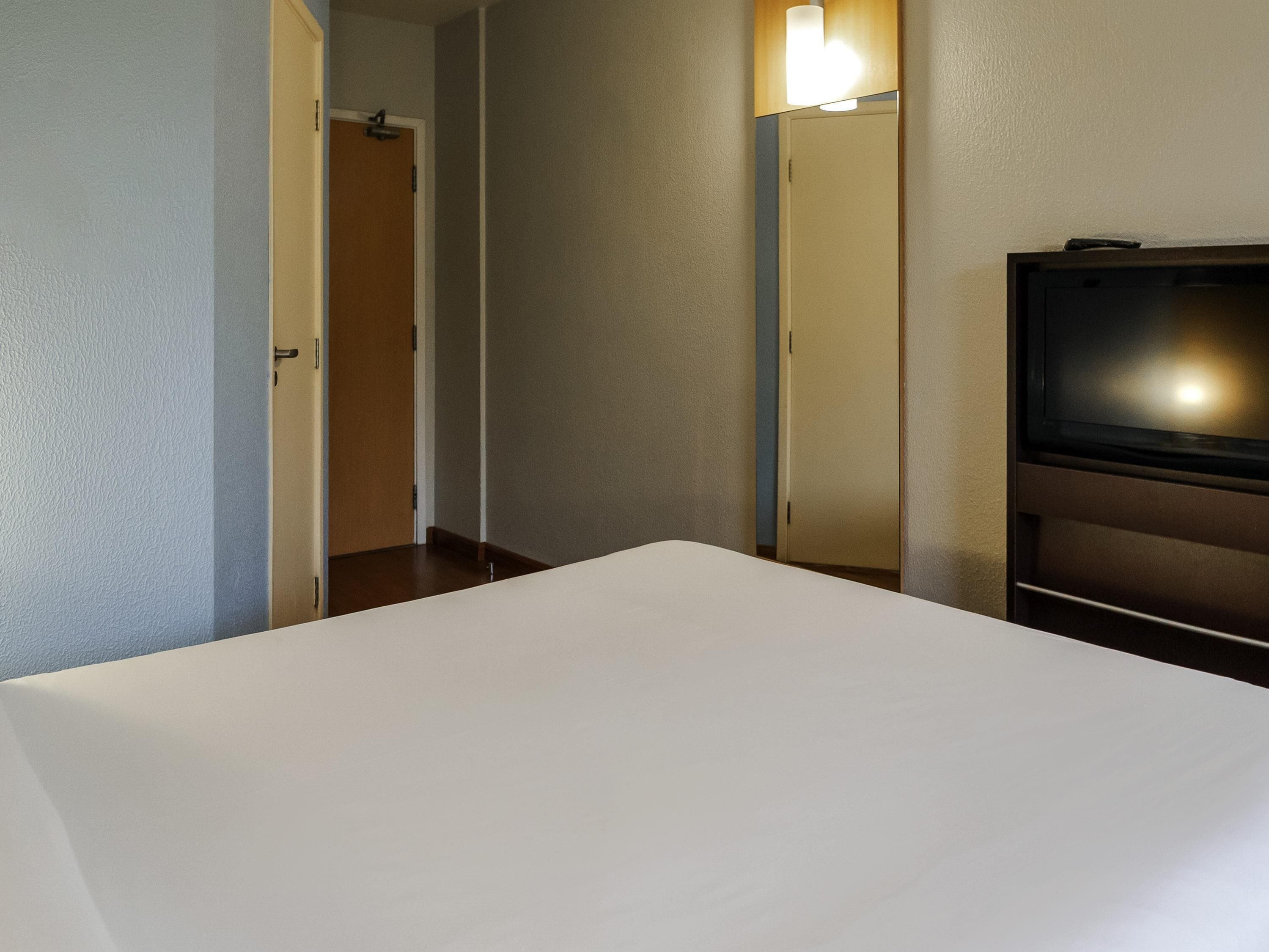 A hotel room with a bed, a television, a door, and a wall-mounted light fixture.
