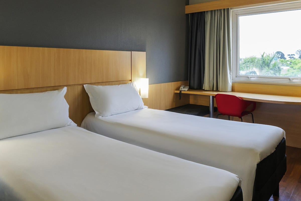 Une chambre d’hôtel avec deux lits, un bureau, une chaise et une fenêtre avec des rideaux.
