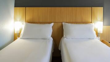 A hotel room with two single beds, wooden headboard, bedside lamps, and a dark wall.