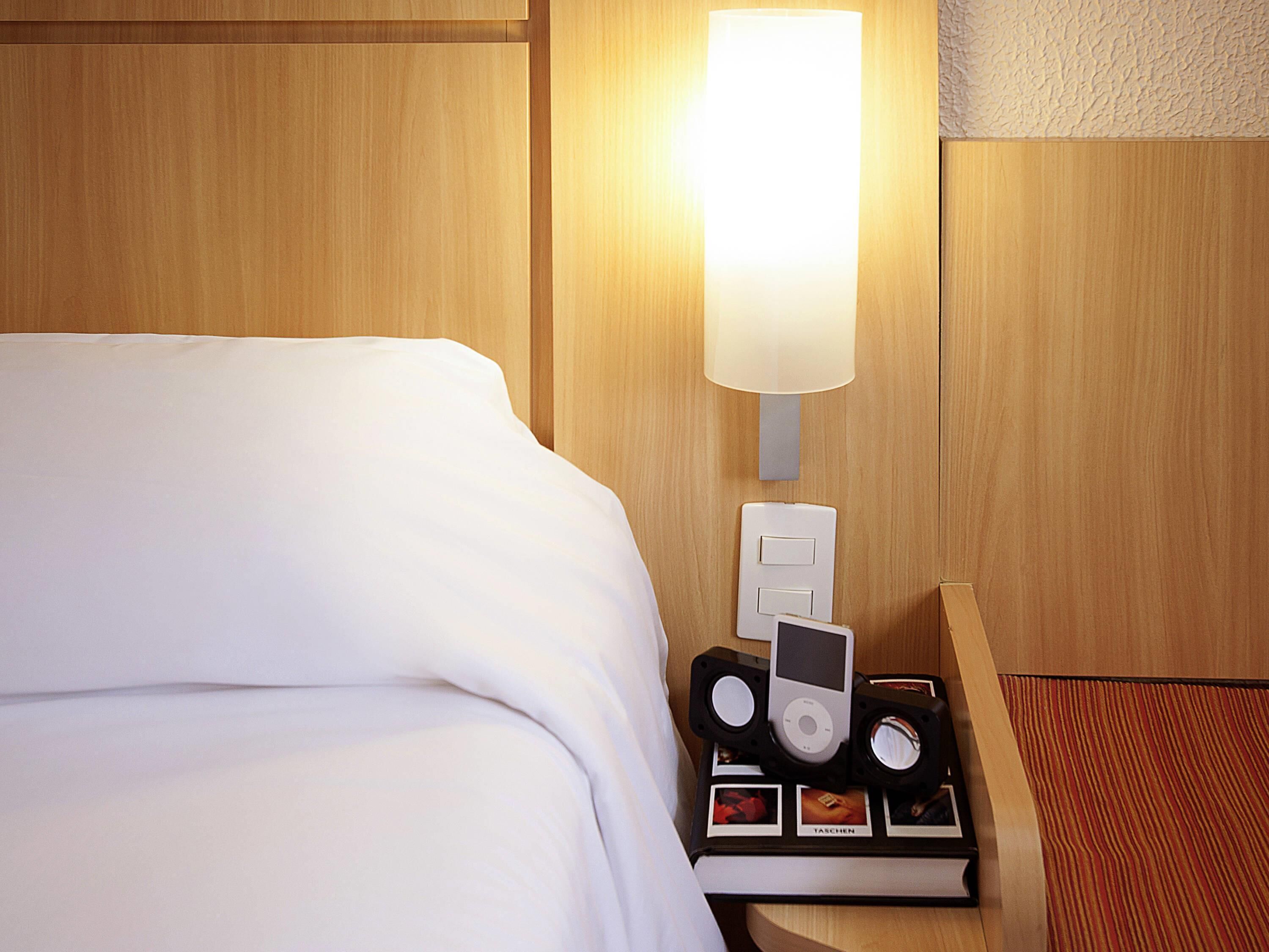 Habitación de hotel con una cama bien hecha, una mesita de noche con lámpara, un reloj despertador y un marco de fotos.