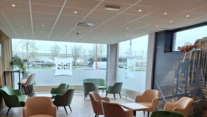 Een moderne zithoek binnen met een combinatie van groene en beige stoelen, een houten vloer en grote ramen met uitzicht op buiten.