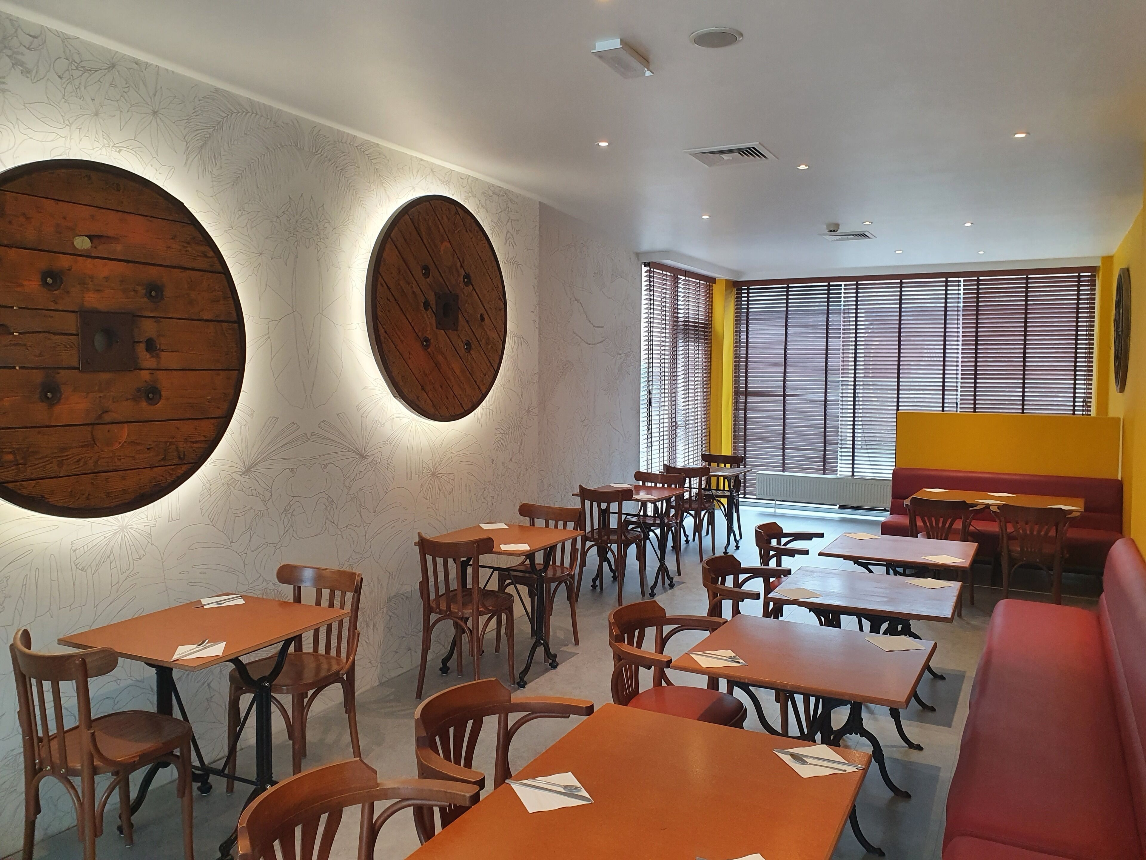 A restaurant with wooden tables and chairs, a red bench, and a large window with blinds.