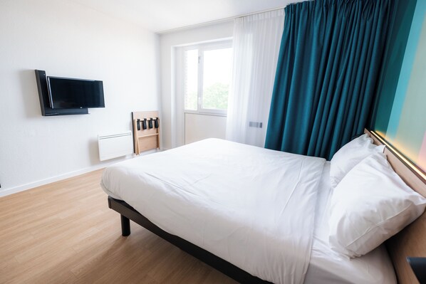 Habitación de hotel con cama, televisor de pantalla plana montado en la pared, ventana con cortinas y suelo de madera.