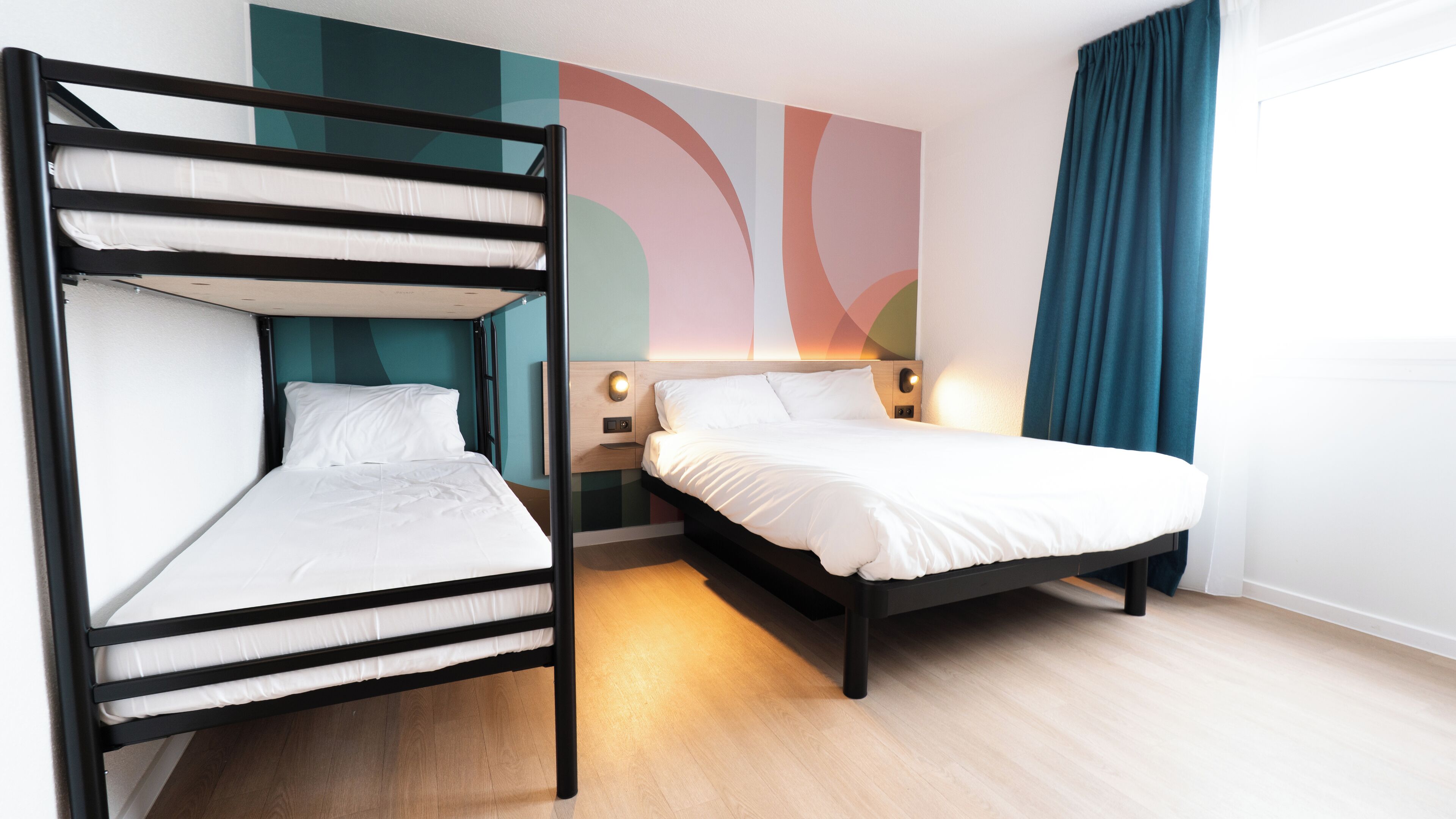 Quarto de hotel com uma cama de beliche, uma cama de solteiro, uma mesa de cabeceira com abajur e uma parede com arte abstrata.
