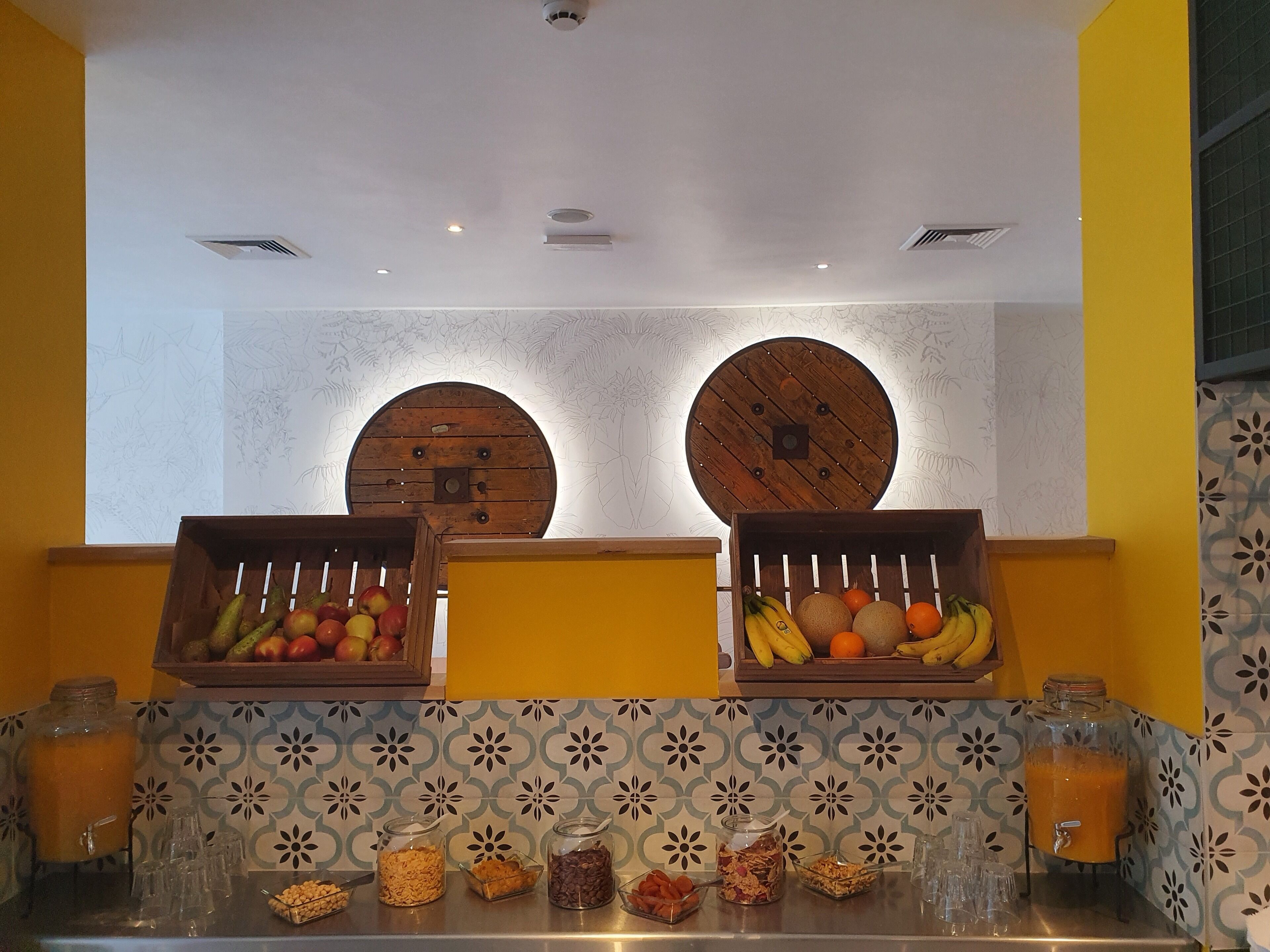 A breakfast buffet with fruit and cereal options, a juice dispenser, and a decorative wall with circular wooden panels.