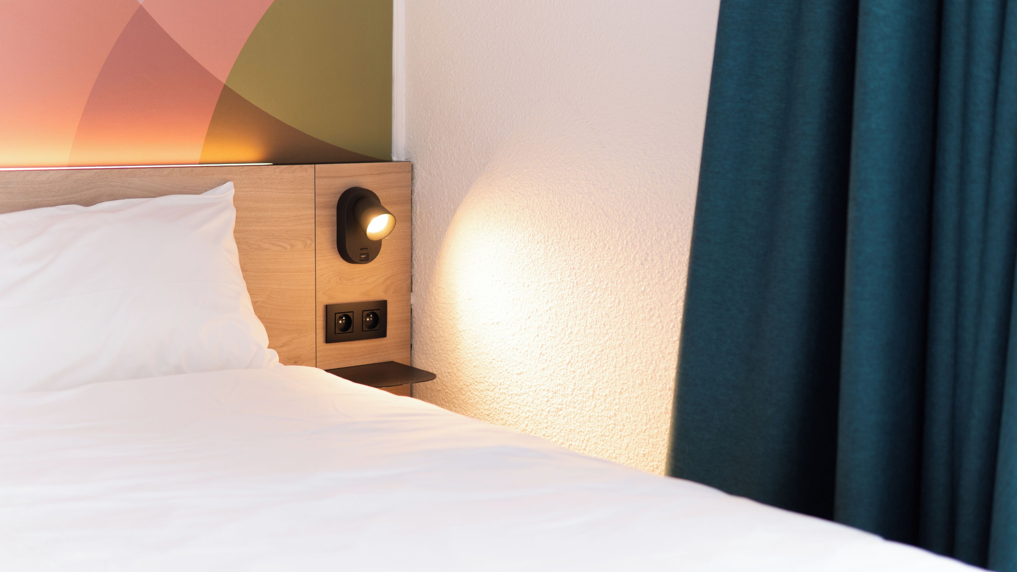 A neatly made bed with white linens, a wooden headboard, a wall-mounted light fixture, and a power outlet.