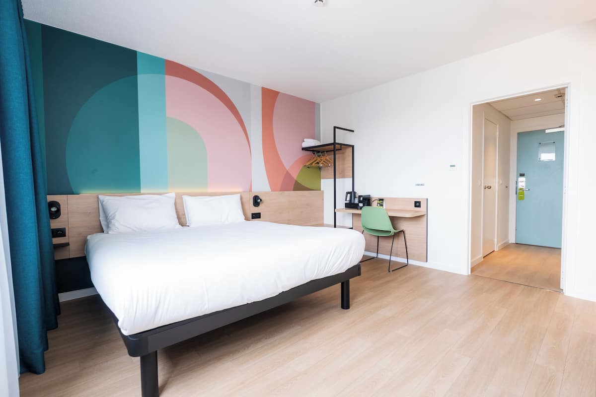 A modern hotel room with a bed, a desk, and a chair. There is a colorful abstract wall art and a door leading to another room.