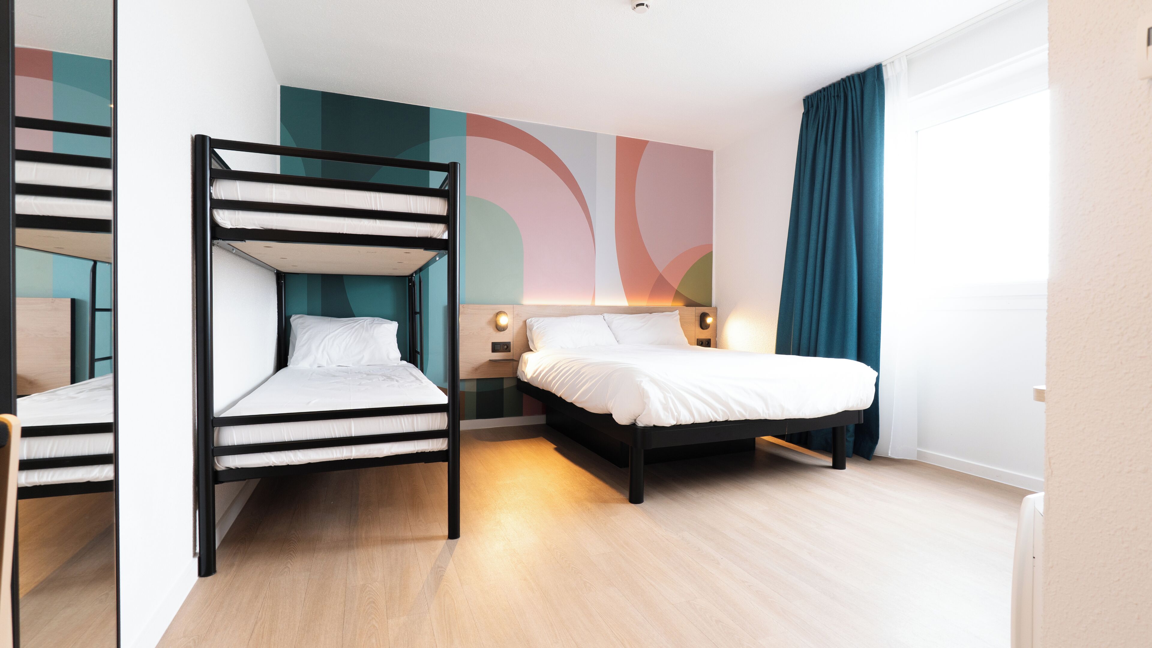 Una habitación de hotel moderna con literas, una cama grande, una pared con arte abstracto y una ventana con cortinas.