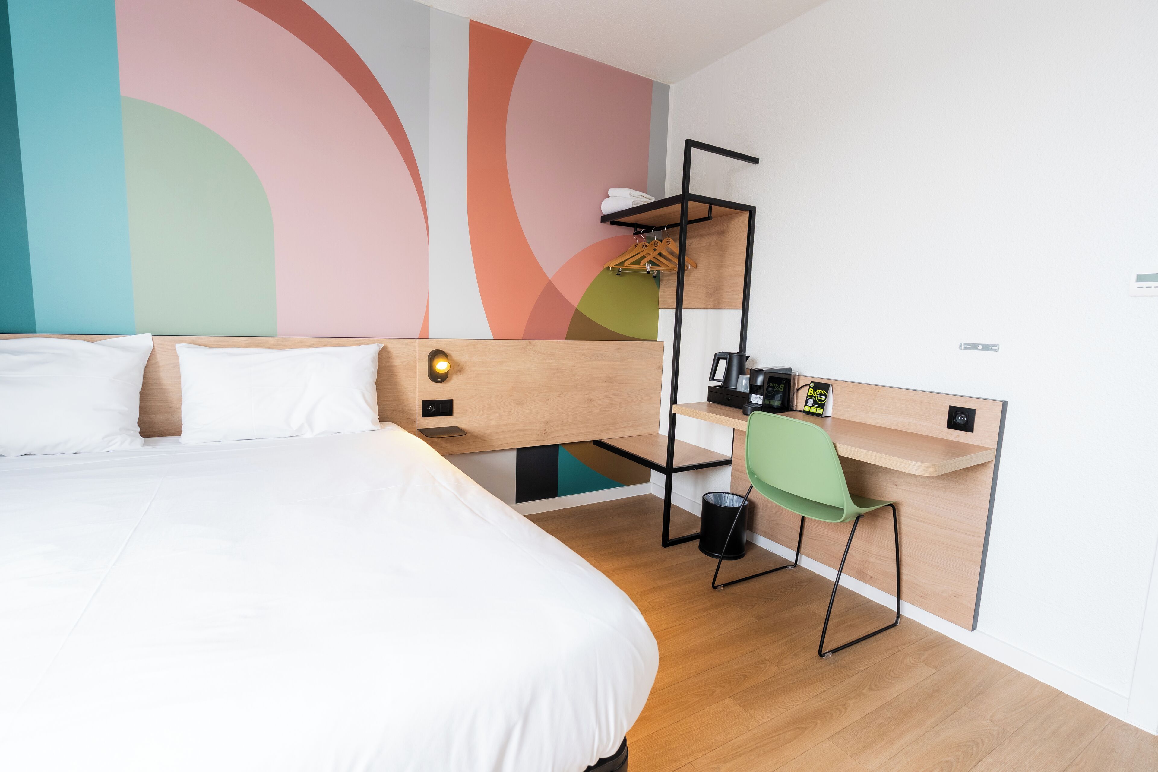 A modern hotel room with a bed, a desk, a chair, and a wall with abstract art.