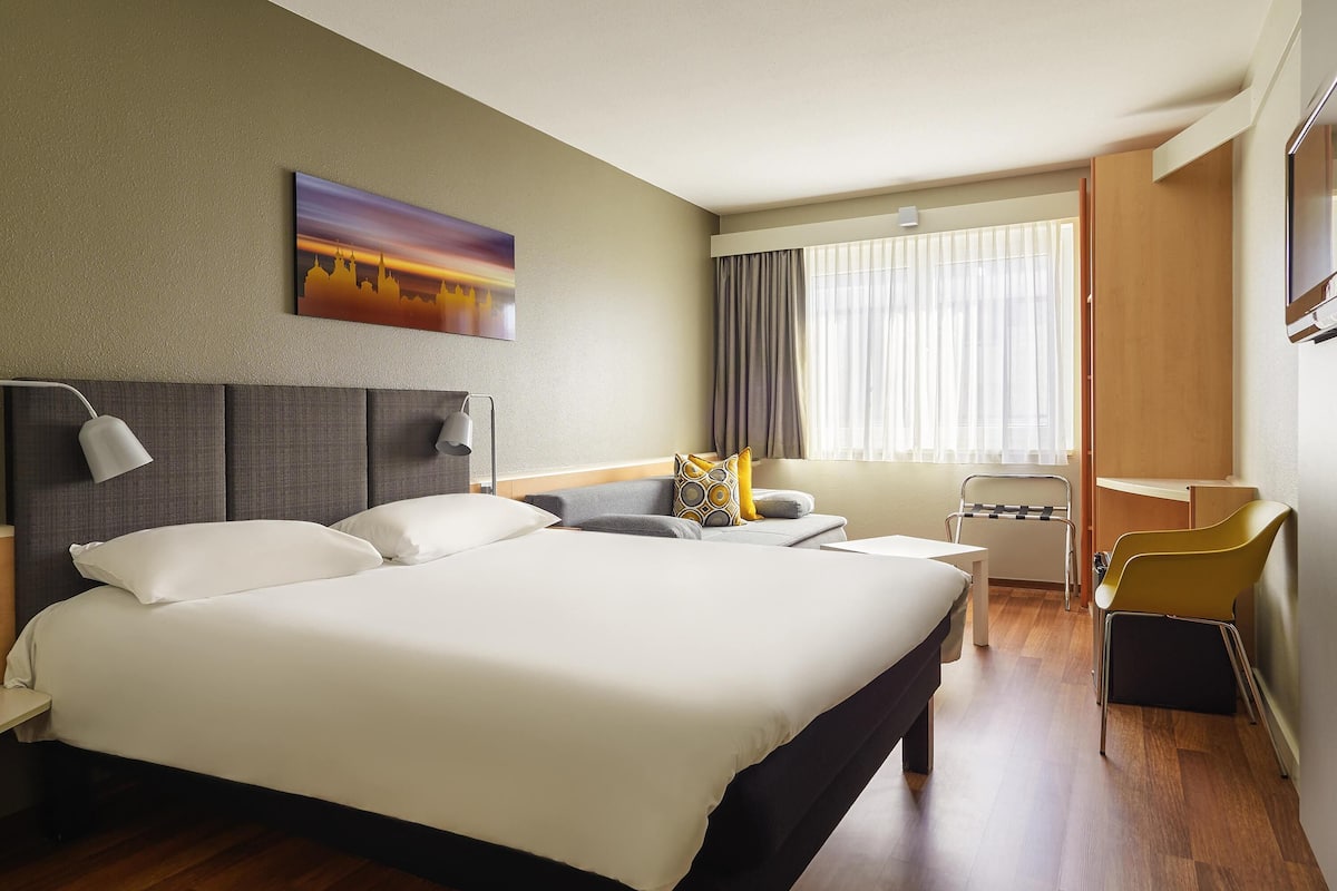 A hotel room with a large bed, a desk, a chair, a TV, and a window with curtains.