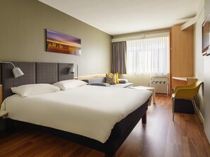 Ein Hotelzimmer mit einem großen Bett, einem Schreibtisch, einem Stuhl, einem Fernseher und einem Vorhangfenster.