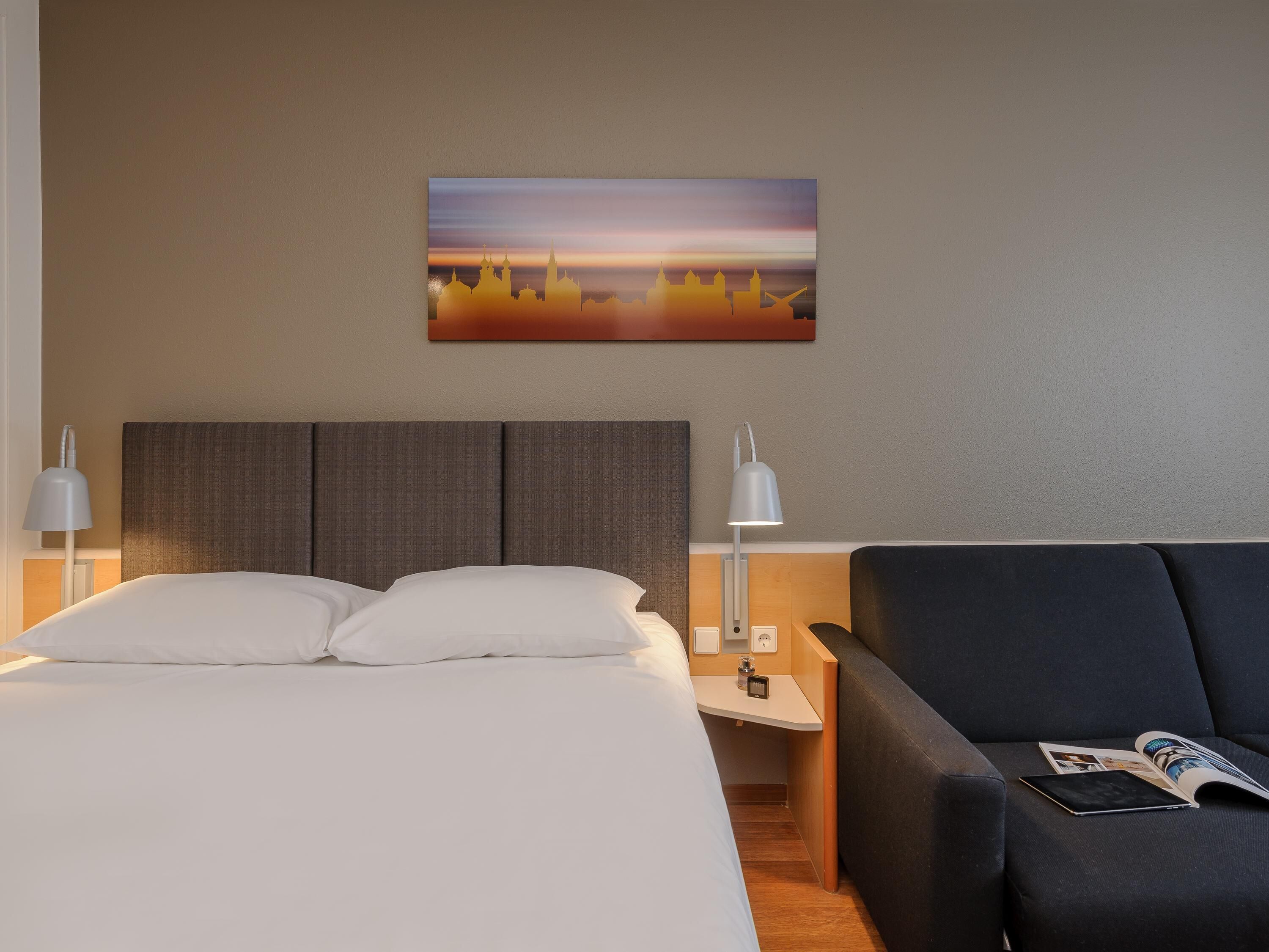 A hotel room with a bed, a nightstand, a lamp, a sofa, and a wall-mounted painting.