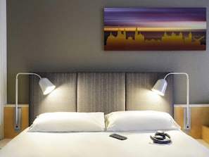 Habitación de hotel con una cama, dos lámparas de noche, un teléfono inteligente y un par de auriculares sobre la cama.