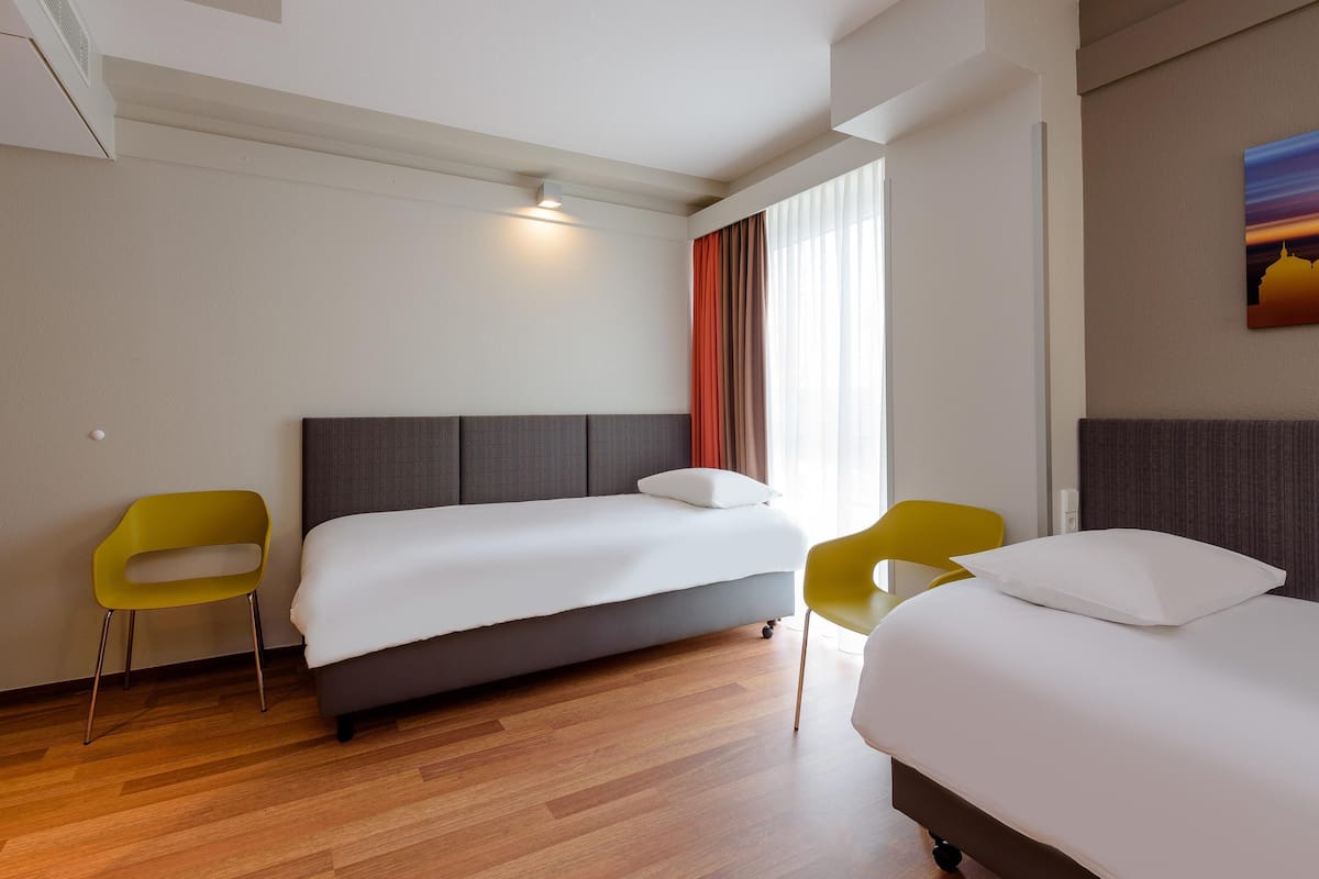 A hotel room with a large bed, two yellow chairs, a window with red curtains, and a painting on the wall.