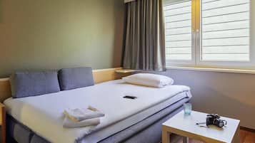 Ett hotellrum med en säng, kuddar, ett nattduksbord med en kamera och ett fönster med persienner.