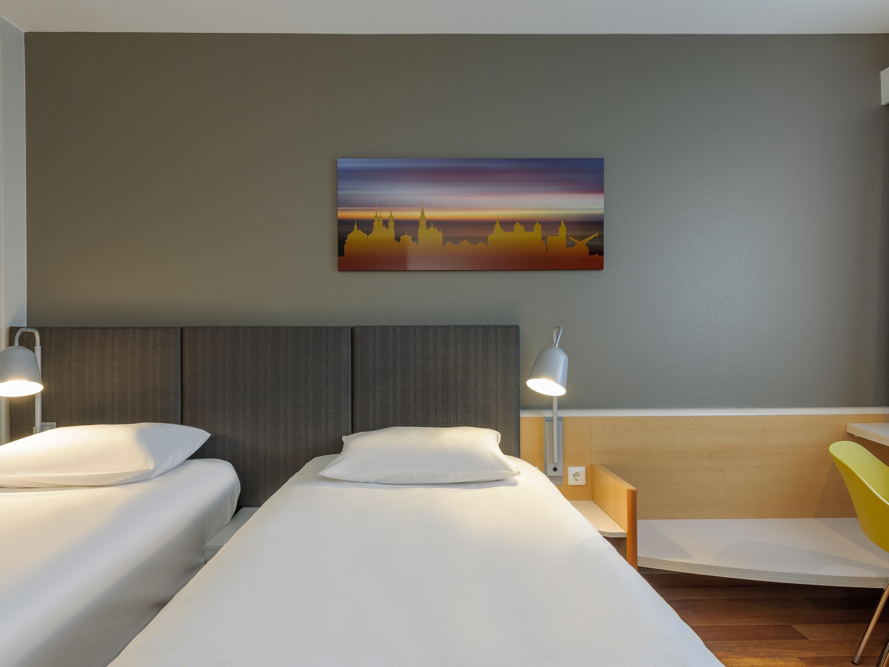 Habitación de hotel con dos camas, un cuadro de un horizonte urbano y un escritorio con una silla.