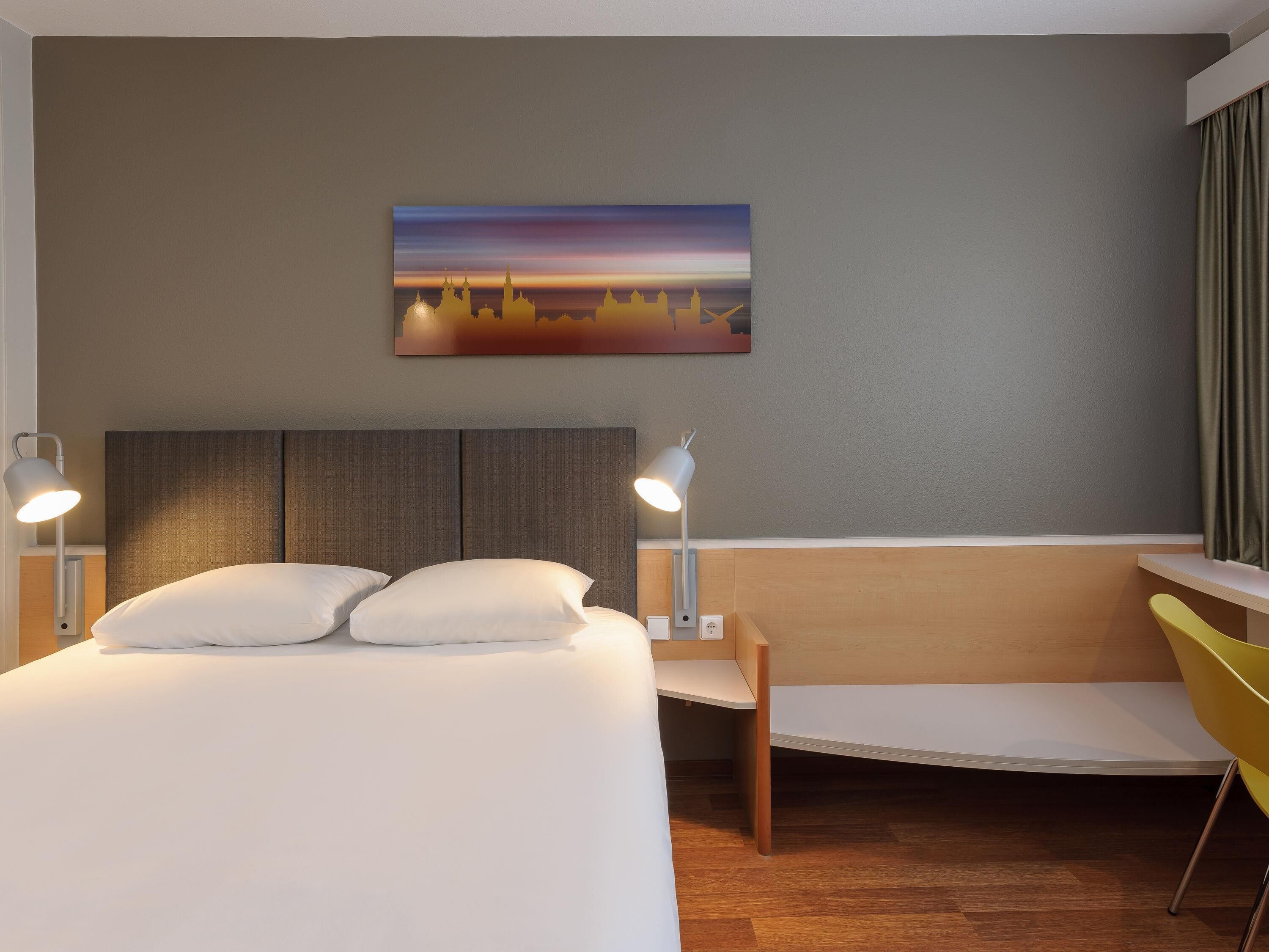 Ett hotellrum med en säng, sänglampor, ett skrivbord, en stol och en tavla på väggen.