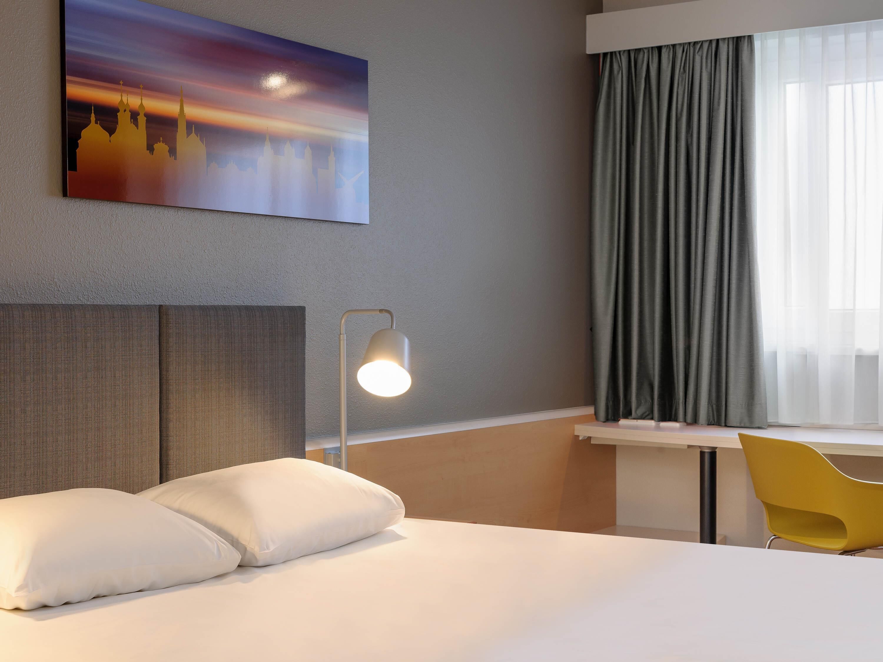 A hotel room with a bed, a desk, a chair, a lamp, and a painting on the wall.