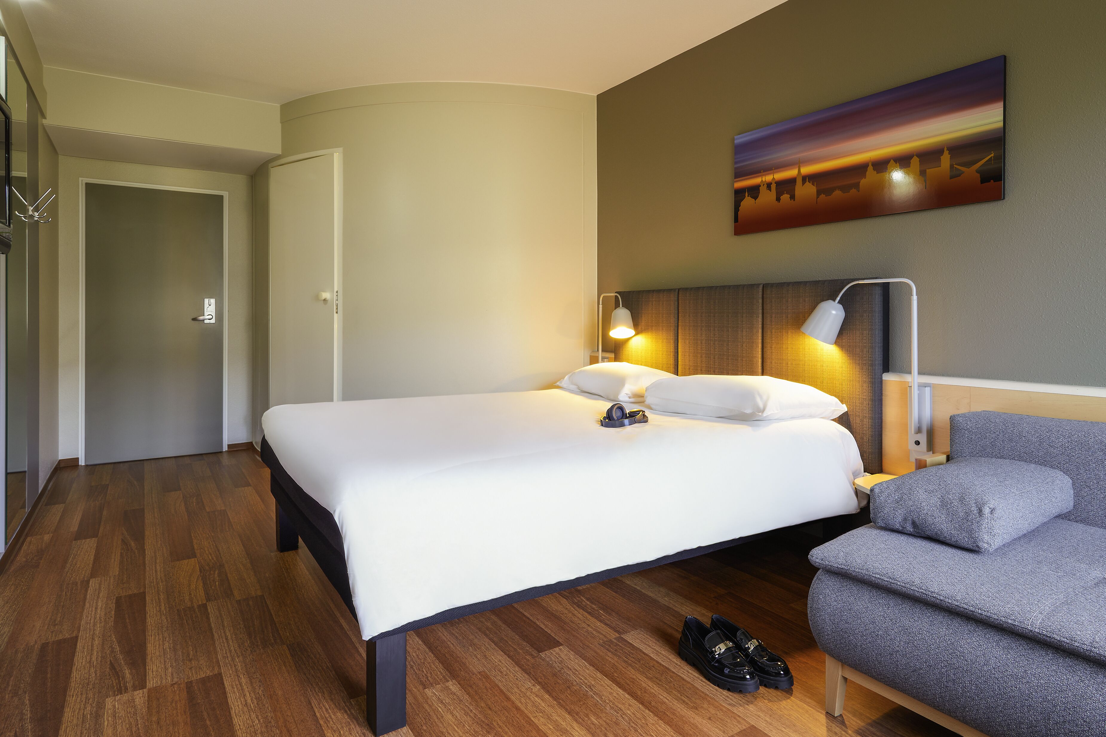 A hotel room with a large bed, a grey sofa, a headboard with a lamp, and a painting on the wall.