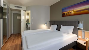 A hotel room with a double bed, a nightstand with a lamp, a painting on the wall, and a bathroom visible through an open door. - ibis Köln Airport (Cologne)