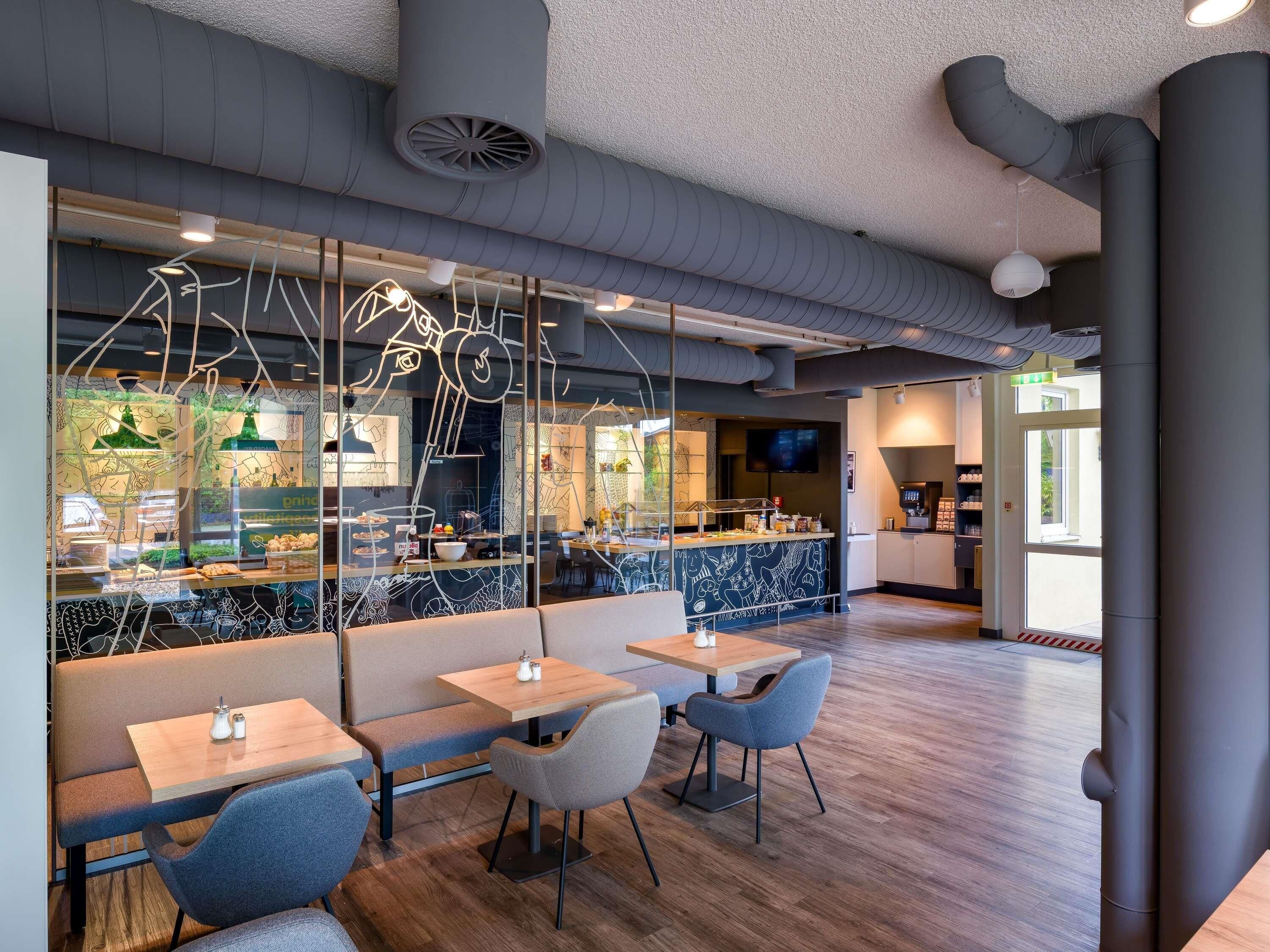A modern restaurant interior with wooden tables and chairs, a counter with a menu display, and a television screen.