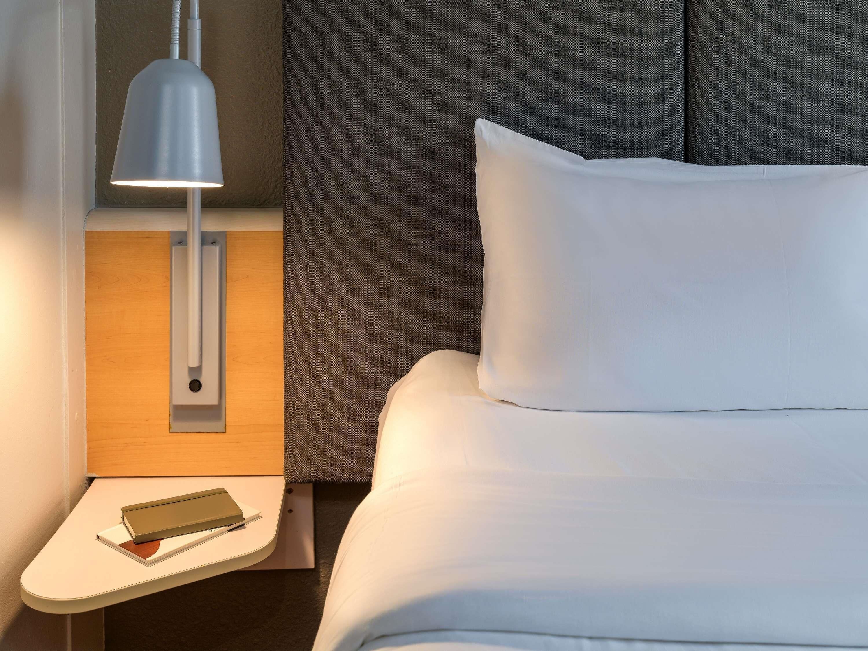 A neatly made bed with white linens, a bedside lamp, a small table with a book, and a textured grey headboard.