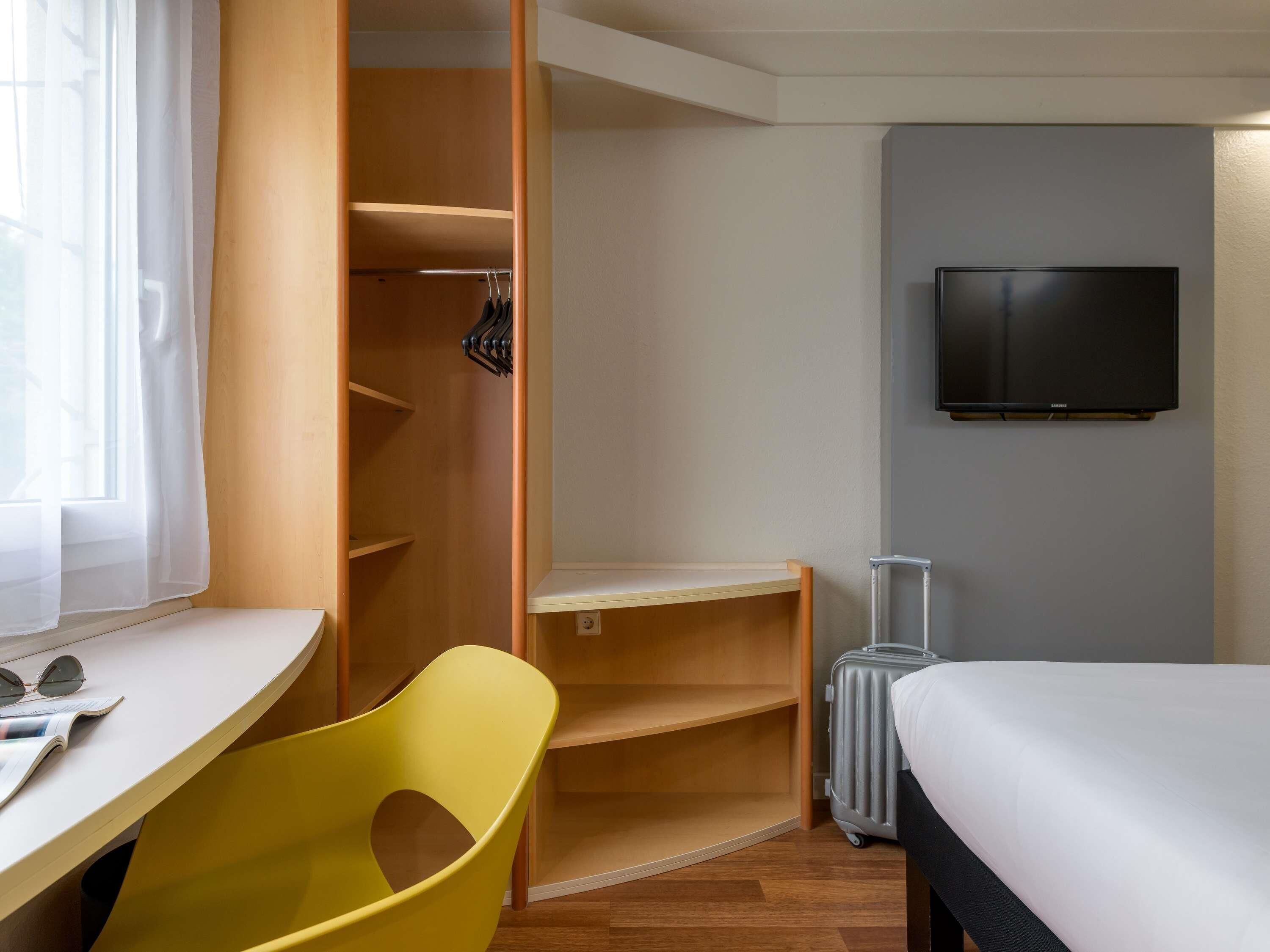 A hotel room with a bed, a desk, a chair, a wardrobe, a suitcase, and a TV.