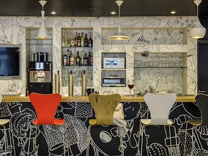 A bar area with a counter, bar stools, a coffee machine, shelves with bottles, and a wall with black and white drawings.