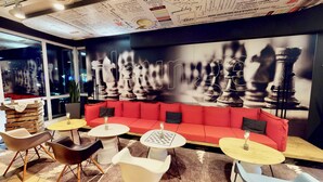 A modern lounge area with a red sofa, multiple tables, and a large chess-themed mural on the wall. - ibis Köln Airport (Cologne)